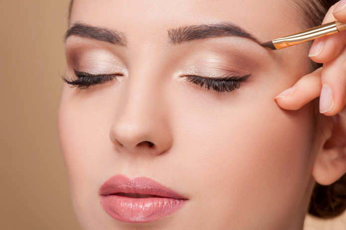 Close up of beautiful face of young woman getting make-up. The artist is applying eyeshadow on her eyebrow with brush. The lady closed eyes with relaxation